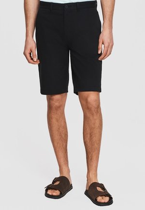 Mężczyzna w czarnych spodenkach do kolan i brązowych sandałach z klamrami, stojący na jednolitym jasnoszarym tle.