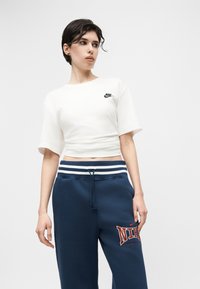 Γυναίκα που στέκεται, φορώντας άσπρο κοντό T-shirt της Nike και μπλε-σκούρα φόρμα της Nike με κορδονάκι και λογότυπο στο μηρό.