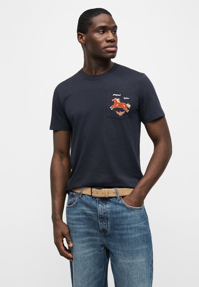 Άνδρας που φοράει μπλε ναυτικό t-shirt με κόκκινο άλογο κεντημένο στην τσέπη του στήθους, μπλε τζιν και μπεζ ζώνη, κοιτάει προς τα πλάγια.