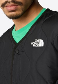 Svart, quilted jakke med v-halsdesign, med knappelukking og logo i hvitt fra The North Face. Grønn skjorte synlig under.