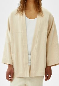 Person som bär en lös beige strukturjacka över en vit skjorta och matchande beige byxor, visad från axlarna till höfterna.