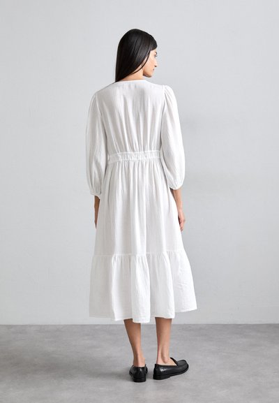 Robe midi blanche à volants avec une taille cintrée, des manches longues bouffantes et un tissu texturé. Associée à des chaussures plates noires.