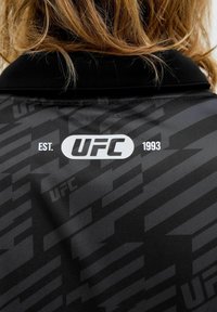 Une personne blonde portant une veste noire UFC avec des motifs géométriques ; le texte blanc sur la veste indique "EST. UFC 1993" au niveau du haut du dos.
