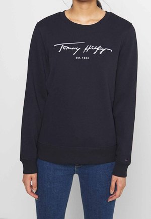 Person iført en sort sweatshirt med lange ærmer med teksten "Tommy Hilfiger est. 1985" foran og blå jeans.