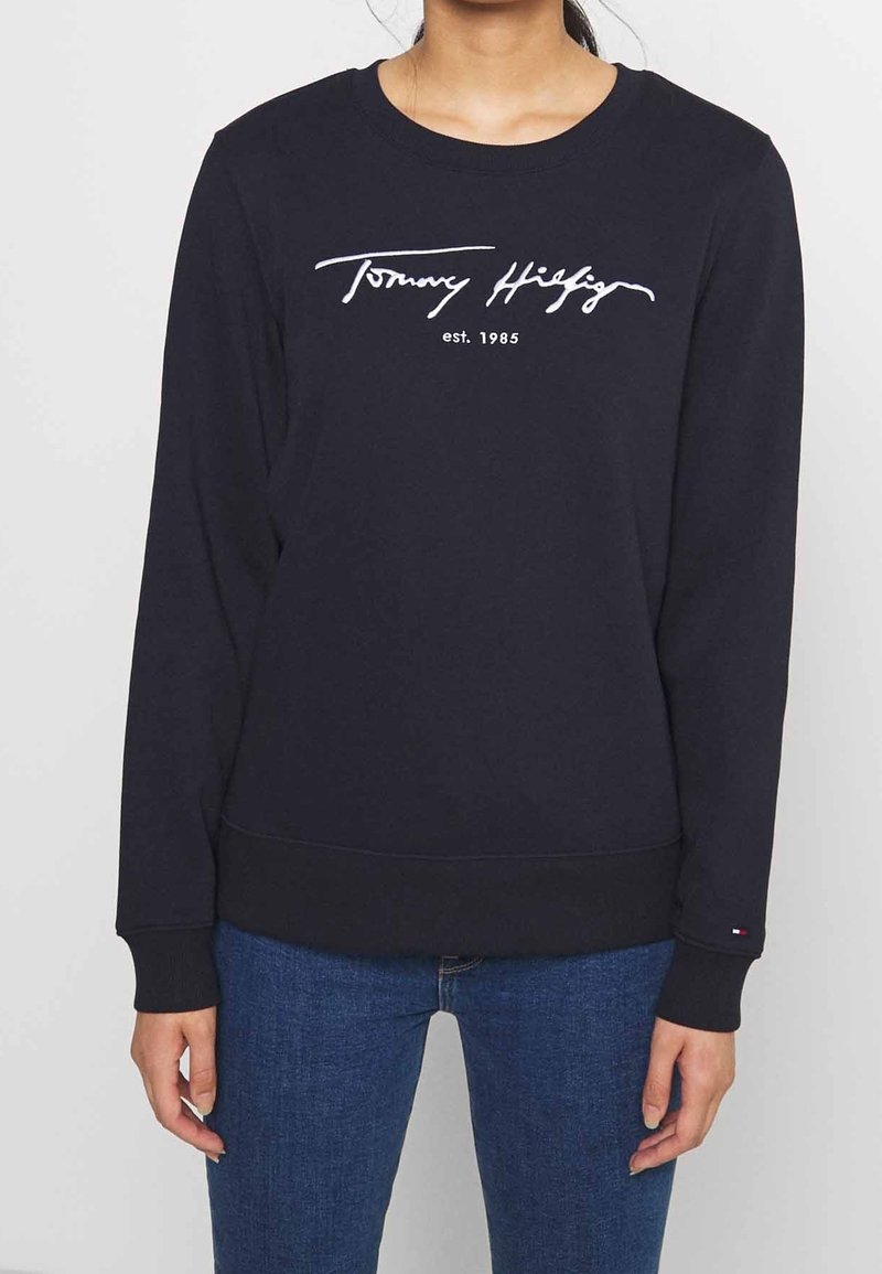 Personne portant un sweat-shirt noir à manches longues avec le texte "Tommy Hilfiger est. 1985" sur le devant et un jean bleu.