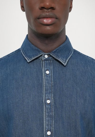 Chemise en denim avec un col boutonné, de couleur bleu clair et boutons blancs, dotée d'un tissu texturé et de détails de couture visibles.