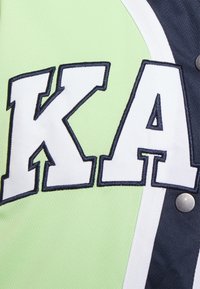 Textureret grøn stof med hvide og marineblå accenter, prydet med store hvide broderede bogstaver "KA" omkranset af marineblå. Sølv trykknaplukning synlig.