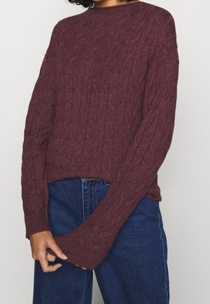 Person trägt einen kastanienbraunen Strickpullover mit langen Ärmeln und blaue hoch taillierte Jeansstoffhosen, die Hände locker vor dem Körper verschränkt.