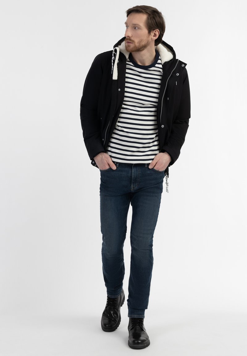 Schwarze Jacke mit weißem Kunstfell-Futter, gestreiftes weißes und marineblaues T-Shirt, blaue Jeans und schwarze Schnürstiefel, Modell steht vor weißem Hintergrund.