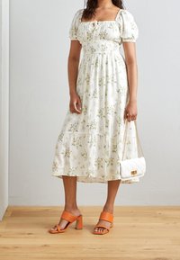 Robe florale blanche avec des manches bouffantes, une taille élastique et une jupe à volants. Associée à des sandales à talons orange et un petit sac à main matelassé blanc.