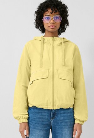 Kobieta o kręconych włosach, nosząca duże fioletowe okulary, jasnżółtą kurtkę z kapturem i niebieskie dżinsy, stojąca na jednolitym tle.