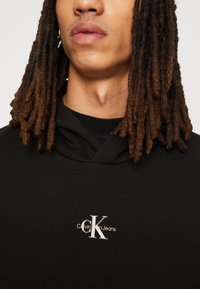 Sudadera con capucha negra de algodón, con un logo de Calvin Klein Jeans bordado en plata y oro en el pecho. Textura suave.