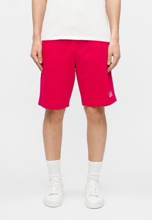 Man wearing bright pink shorts with small white logo on left leg, white crew socks, white sneakers, and a white t-shirt, standing against plain background.