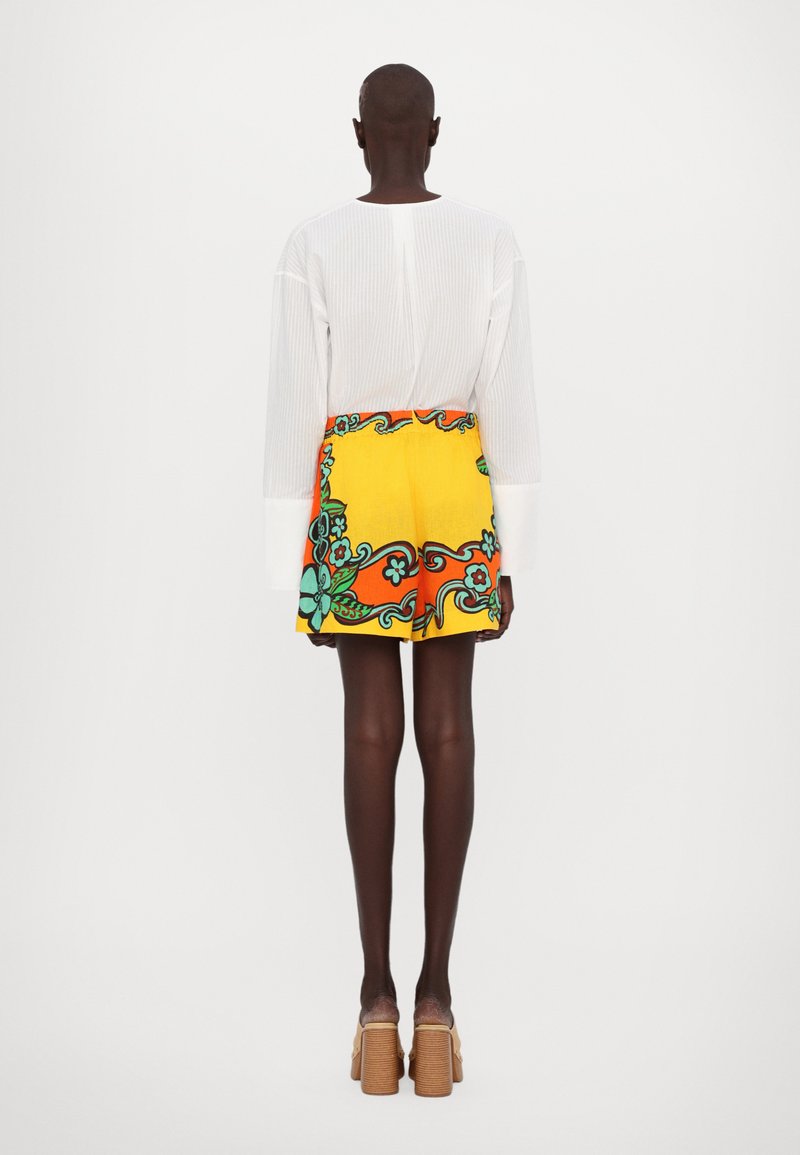 White long-sleeve top paired with a vibrant yellow mini skirt featuring floral patterns in teal and orange, complemented by chunky beige heels.