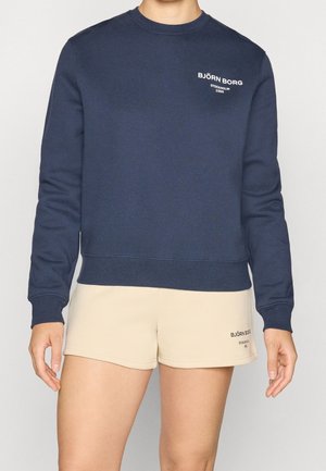 Osoba nosząca granatową bluzę i beżowe krótkie spodenki, oba z tekstowymi logo "BJÖRN BORG STOCKHOLM 1986" na lewej piersi i nodze.