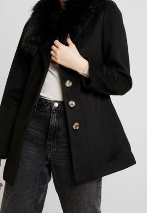 Woman wearing black wool coat with faux fur collar, buttoned front, beige top, and high-waisted black jeans against gray background.