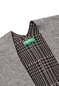 Šedý kardigan s houndstooth vzorem v černé a bílé. Má hluboký výstřih ve tvaru V a zelenou nášivku s nápisem "UNITED COLORS OF BENETTON."