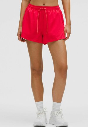 Czerwone spodenki sportowe z elastycznym pasem na sznurek, bocznymi kieszeniami i gładką fakturą. W połączeniu z białym obuwiem sportowym i skarpetkami do kostki.