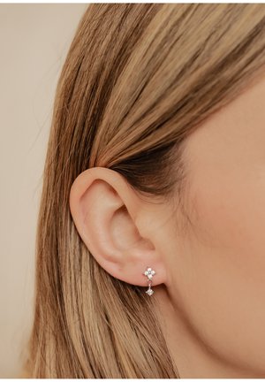 Pendientes de plata con un diseño floral y tres pequeñas piedras preciosas transparentes, colgando justo debajo del lóbulo de la oreja, sobre un fondo claro.