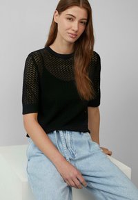 Zwarte gebreide top met een zigzagpatroon en korte mouwen, gecombineerd met lichtblauwe high-waisted jeans. Model zit tegen een neutrale achtergrond.