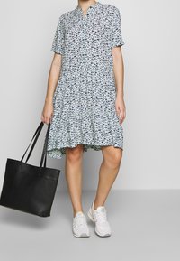 Blaues, kurzärmliges Kleid mit floralem Muster, raffiniertem Taillengürtel und ausgestellt geschnittenem Saum, kombiniert mit einer schwarzen Tragetasche und weißen Sneakers.