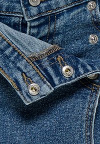 Poignet de veste en denim avec boutons-pression métalliques argentés partiellement déboutonnés, montrant des coutures détaillées et la texture du tissu.