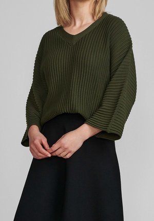 Kobieta w ciemnozielonym swetrze z prążkowanego materiału z dekoltem w szpic i szerokimi rękawami oraz czarnej spódnicy, z luźno złączonymi dłońmi przed sobą.