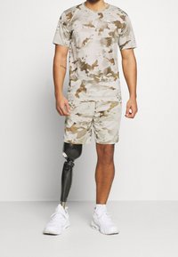 Beżowy zestaw koszulki i szortów w kamuflażu, wykonany z lekkiego materiału. Posiada dopasowany krój, krótkie rękawy i widoczną protezę nogi.