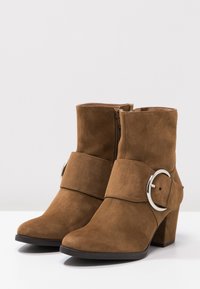 Botas de tobillo de ante marrón con un acento de hebilla plateada redonda, cremallera lateral y un tacón grueso. Textura suave con un diseño de puntera puntiaguda.