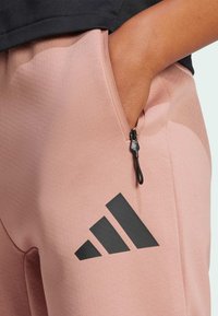 Gaiši rozā sporta bikses ar melnu rāvējslēdzēja kabatu, ar melnu Adidas logotipu sānos un gludu, teksturētu audumu.