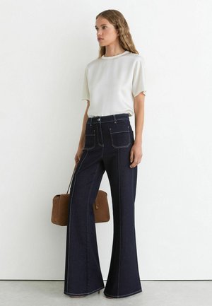 Kvinde med bølget hår iført hvid kortærmet top, mørke vidde jeans og holder en brun håndtaske mod en ensfarvet hvid baggrund.