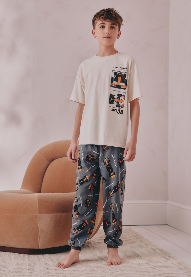 Junge steht barfuß, trägt ein weißes McLaren-T-Shirt und dunkle Hosen mit orange-schwarzen Rennwagenmotiven in einem einfachen Raum.