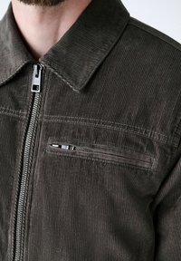 Primer plano de una persona que lleva una chaqueta de pana marrón oscura con una cremallera frontal de metal y un bolsillo en el pecho con cremallera horizontal.