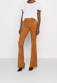 Pantalon évasé en daim marron taille haute, associé à un haut blanc à manches courtes. Chaussures à talons noires avec des clous décoratifs.