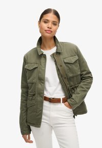 Veste militaire verte avec fermeture éclair et boutons-pression, quatre poches avant, taille et poignets ajustables, portée par-dessus une chemise blanche.