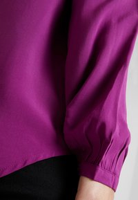 Fuchsia blus med puffärmar och slät textur, som har en subtil fåll och samlade manschetter för en figurnära look.