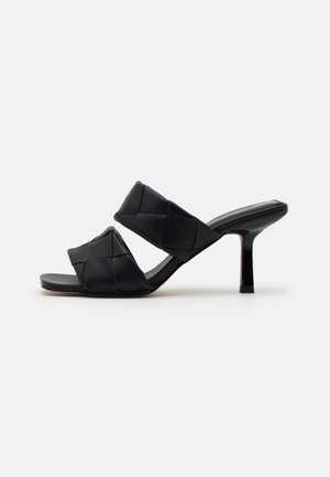 Sort mule-sandal med hæl med to brede, polstrede, quiltede remme og en slank mellemhøj hæl på hvid baggrund.