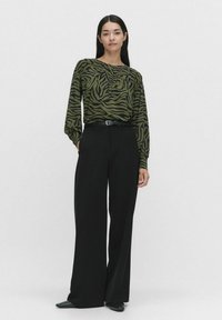 Långärmad blus med zebramönster i grönt och rund halsringning, kombinerad med höga svarta vida byxor och svarta skor.