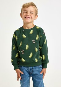 Sweatshirt vert avec impression de gousses de pois et texte "Peas & Love", fabriqué en matière douce, col rond, manches longues et poignets côtelés. Porté avec un jean bleu.