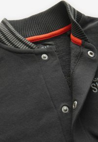 Svart tröja med ribbad krage som har grå och orange detaljer. Funktioner inkluderar tryckknappar och textil med struktur.