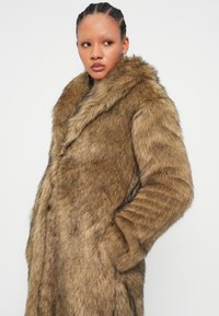 Καφέ ψεύτικο παλτό από γούνα με απαλή υφή, διαθέτοντας φαρδύ γιακά, πλαϊνές τσέπες και κάθετες ρίγες στα μανίκια.