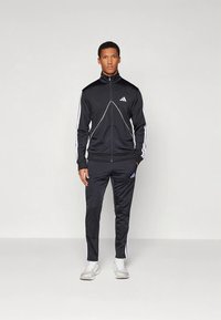 Μαύρη αθλητική φόρμα από λείο ύφασμα, με λευκές ρίγες στα πλάγια, φερμουάρ και έμβλημα στο στήθος και το παντελόνι.