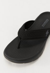Black flip flop made of textured rubber, featuring a woven strap and a cushioned footbed with a wavy pattern. Logo embossed on the side.