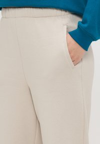 Lichtbeige joggers met een elastische tailleband en zijzakken. De stof heeft een gladde textuur met subtiele stikdetails.
