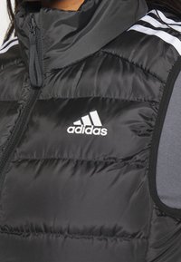 Svart vadderad väst med hög krage, utrustad med dragkedja fram och vit Adidas-logotyp. Strukturerad tyg med horisontella sömmar och grå accenter.