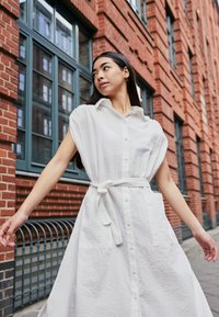 Vestido branco de manga curta com frente abotoada, cintura com cordão e tecido texturizado com suaves riscas verticais. Duas grandes bolsos frontais.