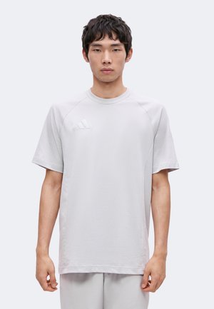 Junger Mann, der ein schlichtes hellgraues kurzärmeliges T-Shirt mit Adidas-Logo auf der oberen linken Brust trägt, steht vor einem hellgrauen Hintergrund.