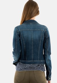 Chaqueta de mezclilla en azul oscuro, con un diseño ajustado, costuras prominentes y una longitud ligeramente recortada. Lleva puesta una camisa de rayas azul y blanca.