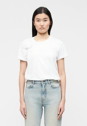 Frau mit glatten schwarzen Haaren, die ein einfaches weißes kurzärmeliges Hemd und hellblaue, hoch taillierte Jeans trägt, steht vor einem weißen Hintergrund.