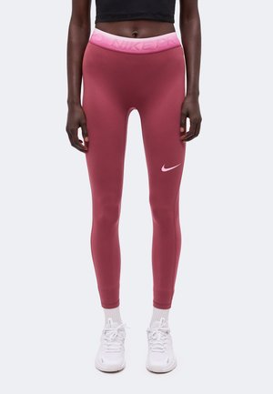 Osoba nosiaca ružové legíny Nike s vysokým pásom, biele ponožky a biele Nike tenisky, stojaca pred jednoduchým svetlým pozadím.